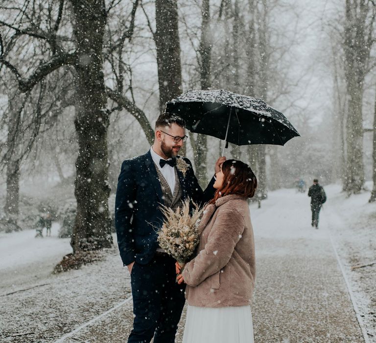 Bride in fur coat holding dried flower bouquet looks up stands under umbrella in the snow with groom in navy suit and tartan waistcoat