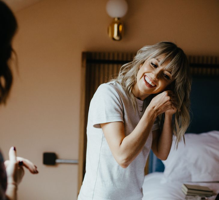 White, blonde bride with fringe and wavy hair putting in earrings and smiling