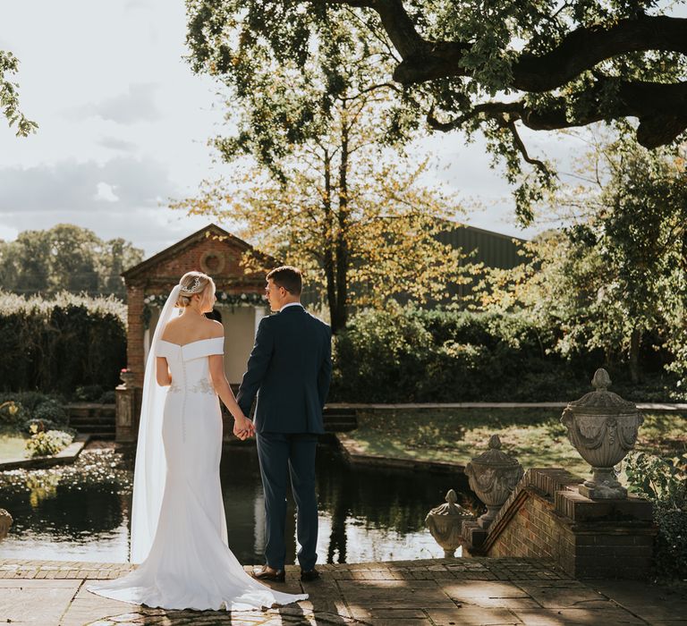 Bride & groom hold hands beside pond in the sunshine