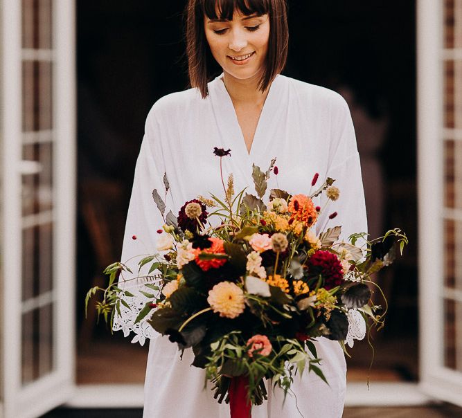 Bride with bob and fringe on the wedding morning in a white getting ready robe holding her colourful bouquet 