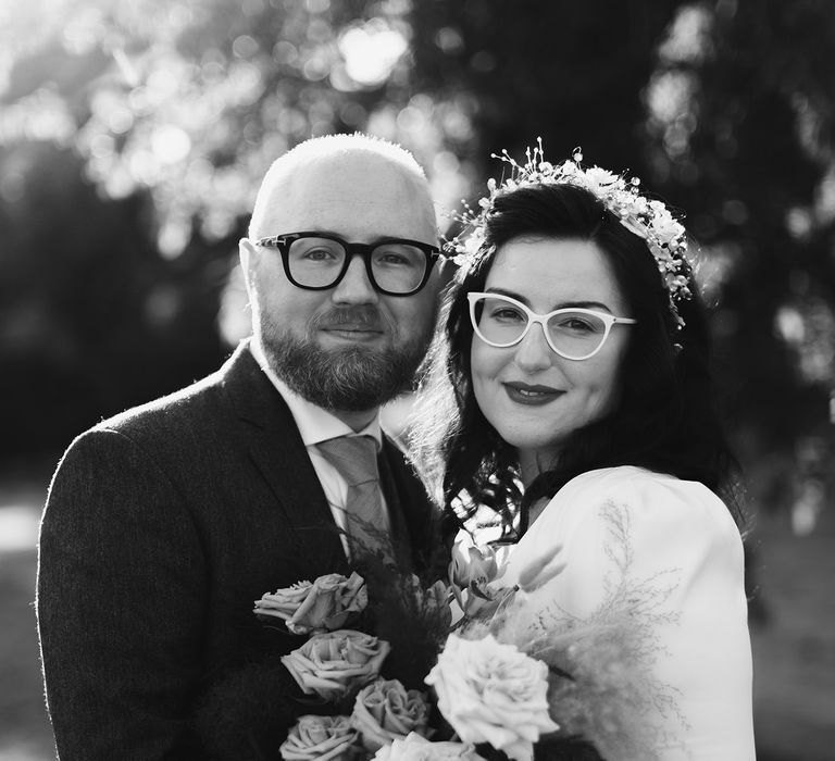 Groom in navy suit and yellow tie stands with bride in white cat eye glasses and bridal headband holding white and pink rose and pampas grass bouquet