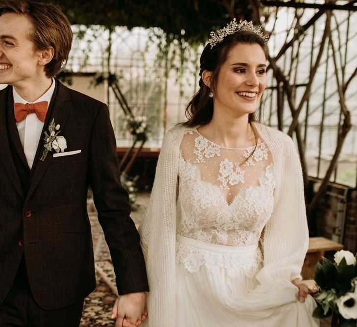 Groom in a three-piece suit with orange bow tie holding hands with his bride in a lace and tulle dress with wool wrap and crown