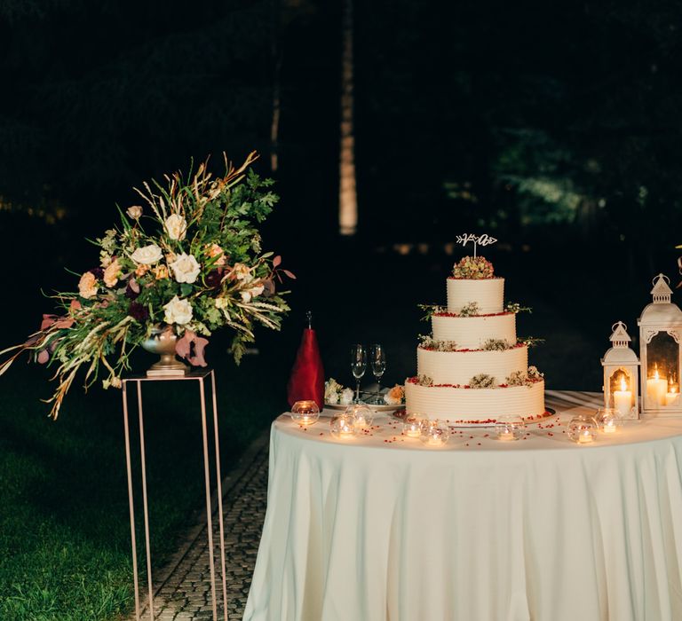 Huge white iced four tier wedding cake at night, on a table with candle lanterns and floral bouquets