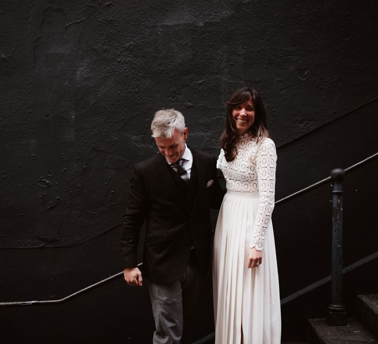 Bride in long sleeved lace top Self Portrait wedding dress stands with groom in brown woollen blazer and grey trousers on steps by black wall in Bristol