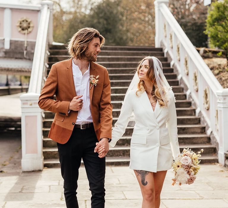 Bride in a blazer dress and appliqué veil holding her grooms hand black chinos and rust blazer in Chelsea 