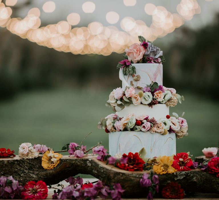 White, blue and pink three tiered wedding cake with multicoloured rose and foliage decor on wooden bark-lined stand in fairy-lit olive grove at enchanted forest wedding in Italy