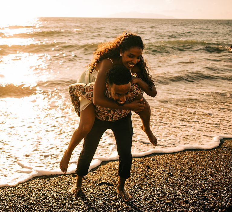 Groom carries bride on his back as the sea shines in the background 