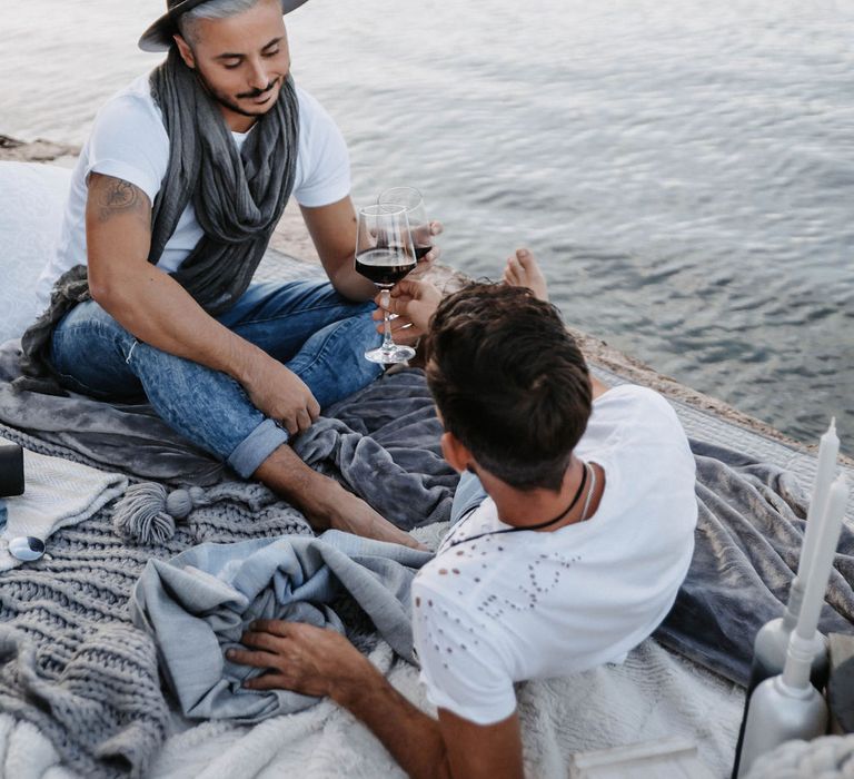 Grooms lay on blankets beside the sea as they drink red wine