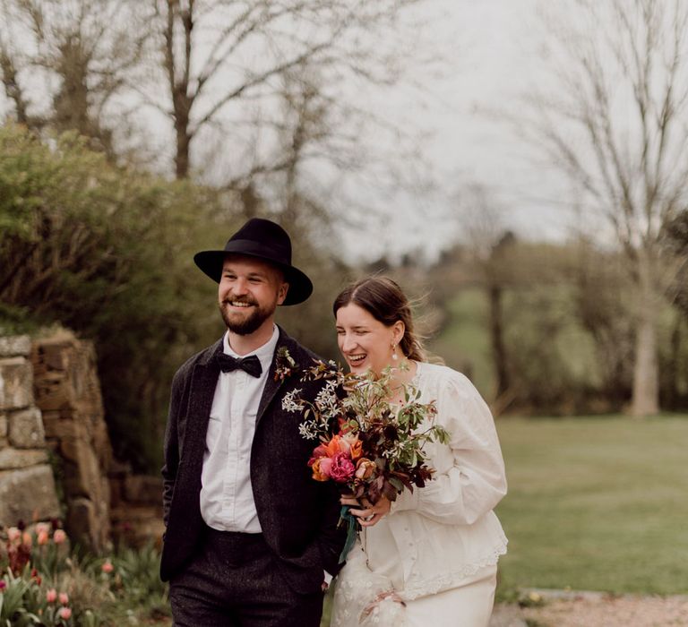 Smiling bride in Charlie Brear wedding dress and chapel length daisy applique veil holding multicoloured wedding bouquet walks arm-in-arm with groom in grey tweed suit and black fedora at garden party wedding in Devon