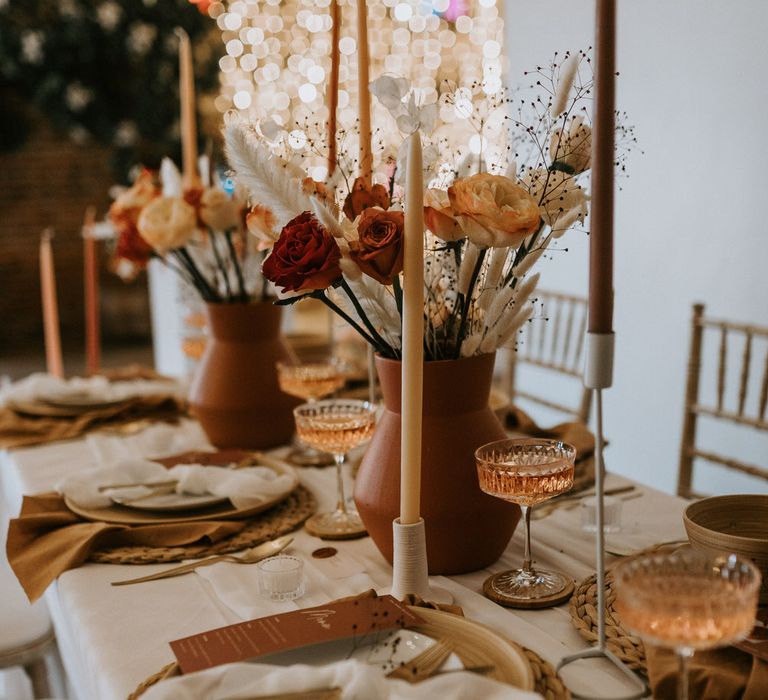 Neutral tablescape with tapered candles, a terracotta vase of flowers, rose champagne in crystal cut coupes, gold cutlery and white plates