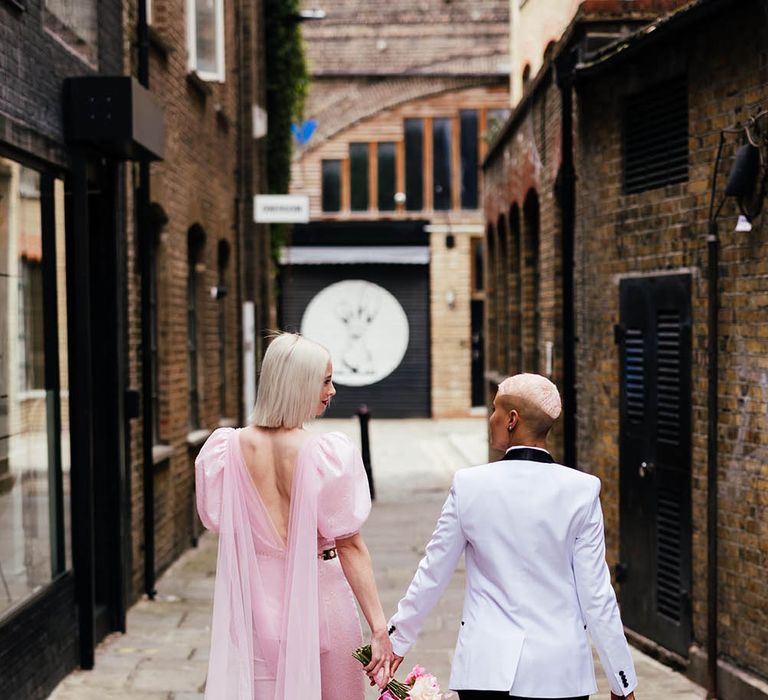 Bride in a custom-made pink jumpsuit with slim leg trousers and puff sleeves holding hands with a her bride in a white tuxedo jacket 