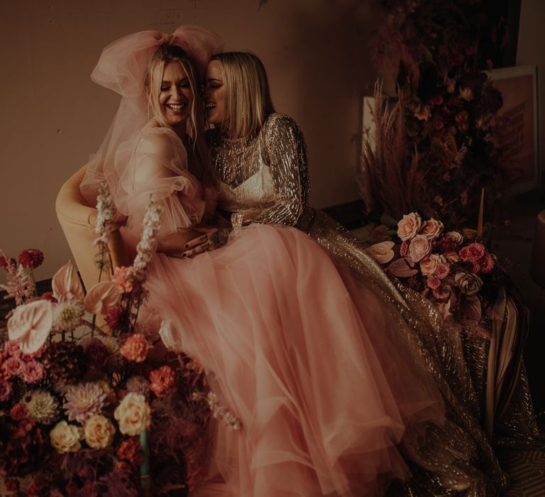 A lesbian couple sit together laughing. One wears a tulle pink dress with a large pink bow in her hair and the other wears a gold embellished dress. Image by Esme Whiteside.