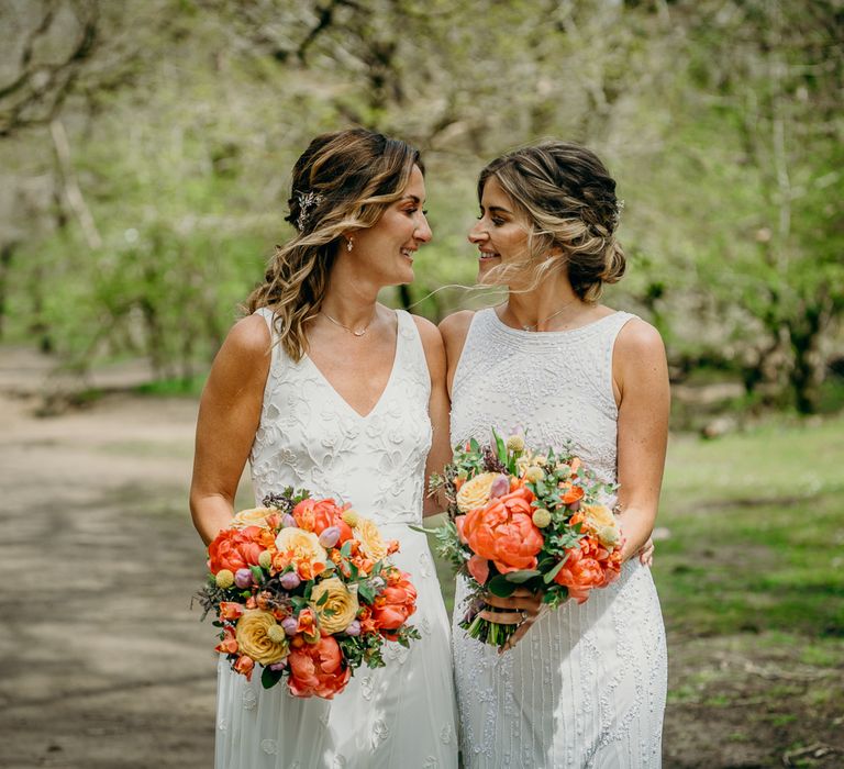 Brides look lovingly at one another whilst holding bright floral bouquets