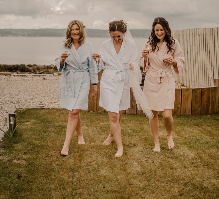 Bride in veil and two guests all in short dressing gowns stand smiling in the garden all holding sparkling wine