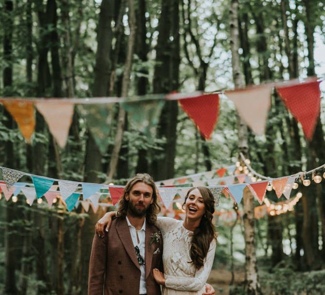 Bride in a lace boho wedding dress with her arm around her husband in a brown suit standing in the woods decorated with colourful bunting decor 