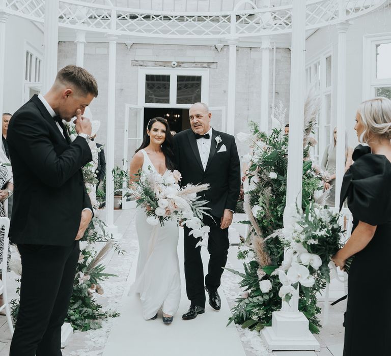 Groom rubs eyes as bride in white Pronovias gown holding white, pink and green wedding bouquet walks down the aisle in the glass conservatory at Came House Dorset arm in arm with man in tuxedo