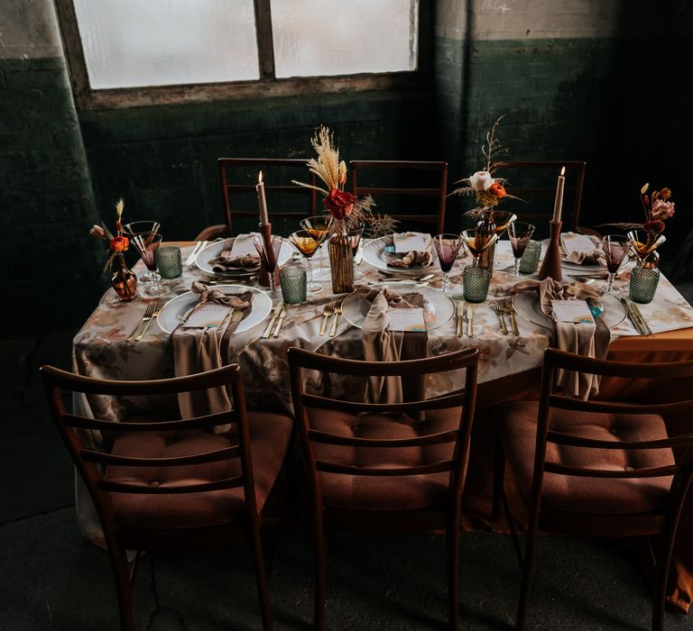 70s wedding table decor with patterned table cloth, gold cutlery and rimmed charger plates, coloured glassware and dried and fresh flowers in vessels 