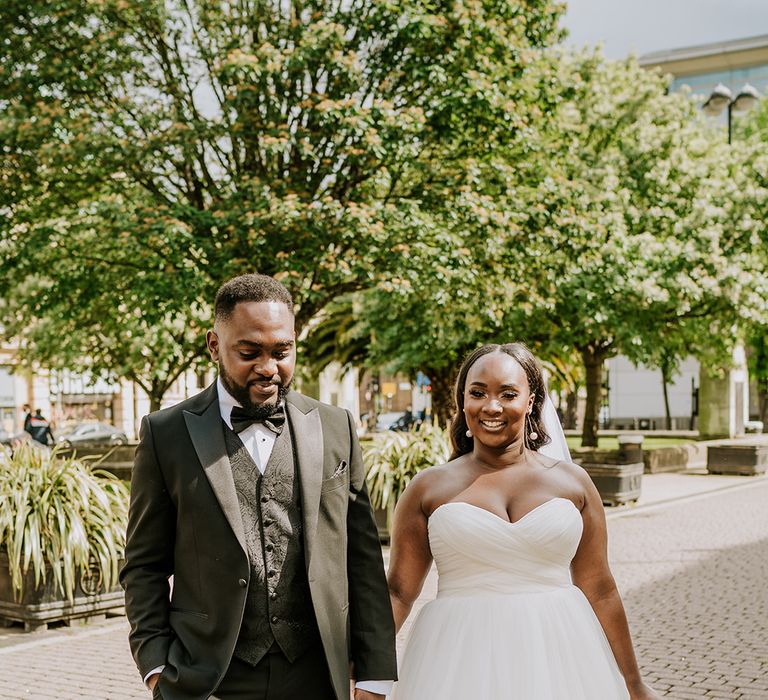 Bride & groom walk with one another outdoors on their wedding day as the sun shines and they smile 