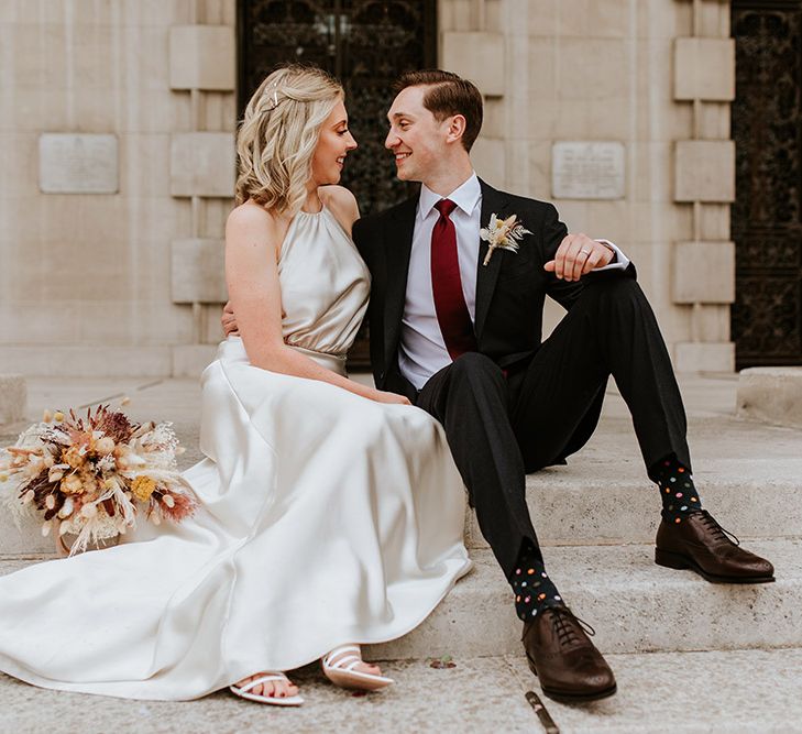 Bride and groom portrait outside Leeds Civic hall with bride in a champagne colour wedding dress sitting next to her groom in a black suit and burgundy bow tie 