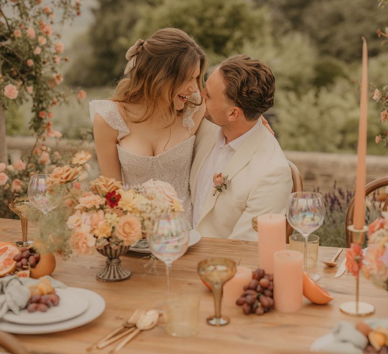 Bride in a sparkly Made With Love wedding dress sitting on her grooms lap in a beige suit at their outdoor wedding reception decorated with pastel orange flowers 