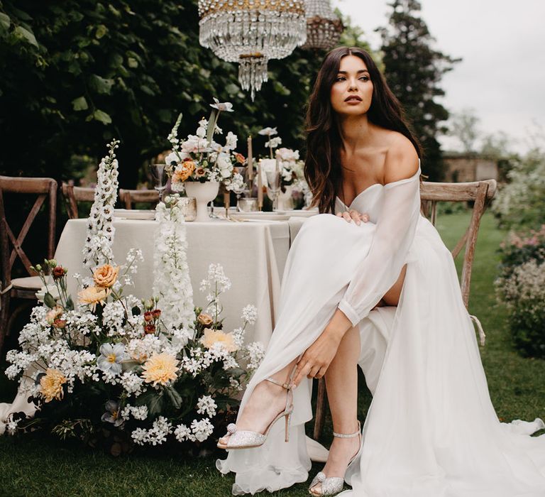 Bride in an off the shoulder chiffon wedding dress with long sleeves and front split adjusting the straps on her silver wedding shoes 