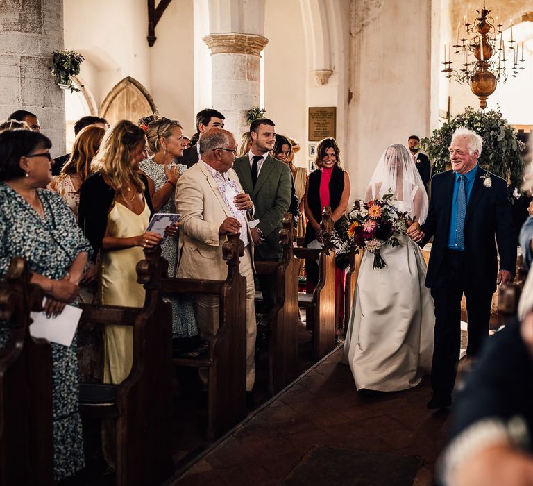 Bride walks down the aisle wearing veil carrying floral bouquet