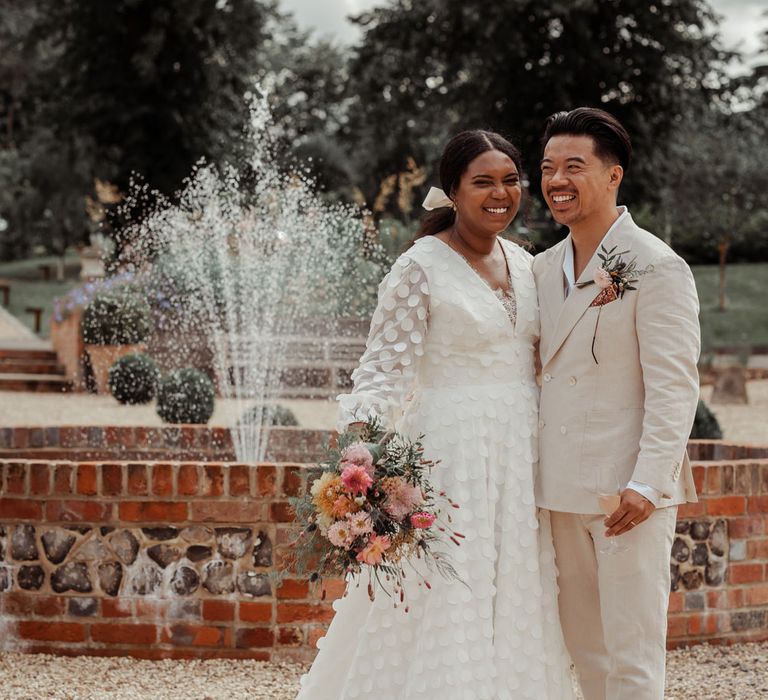 Bride in white long mesh sleeve spotted Vagabond wedding dress and bridal hair bow holding mixed bridal bouquet stands with groom in white shawl collared shirt and double breasted linen suit with brown shoes next to fountain outside in gardens at Wasing Park wedding