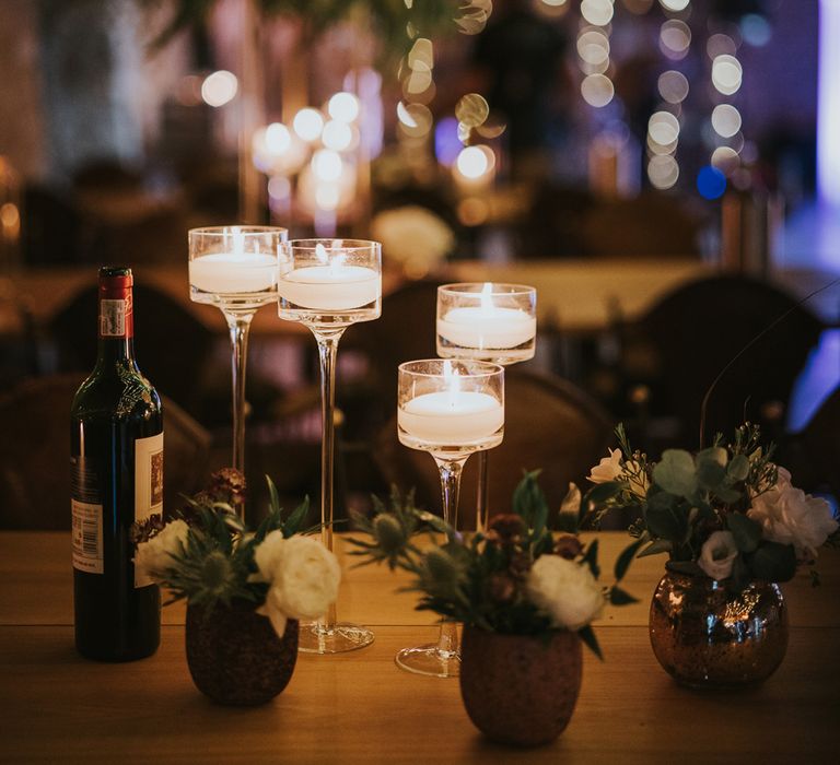 Glass long stem candle holders next to flowers in small gold vases and bottle red wine in fairy lit room at Tythe Barn wedding with barn wedding flowers