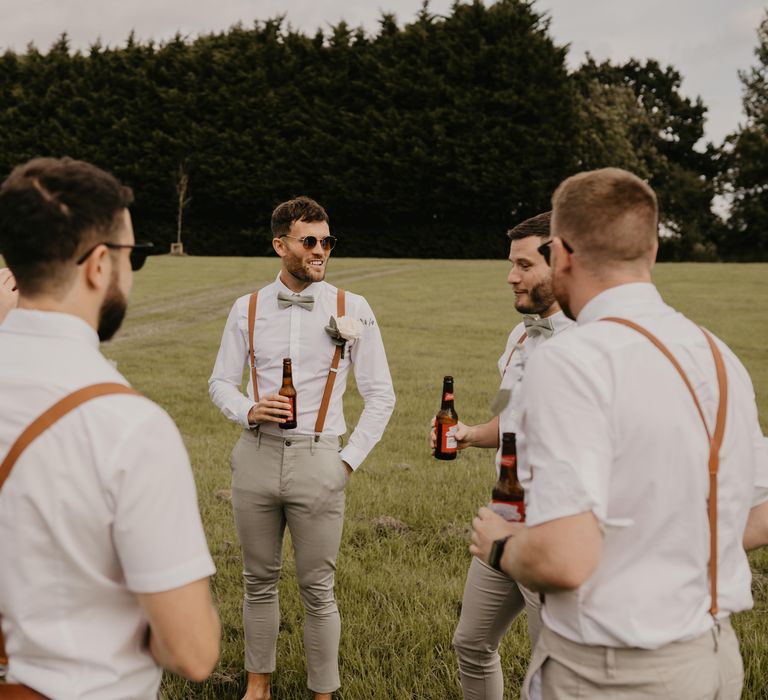 Groom & his groomsmen stand outdoors as they drink and groomsmen wear shirts with brown braces | Mark Bamforth Photography