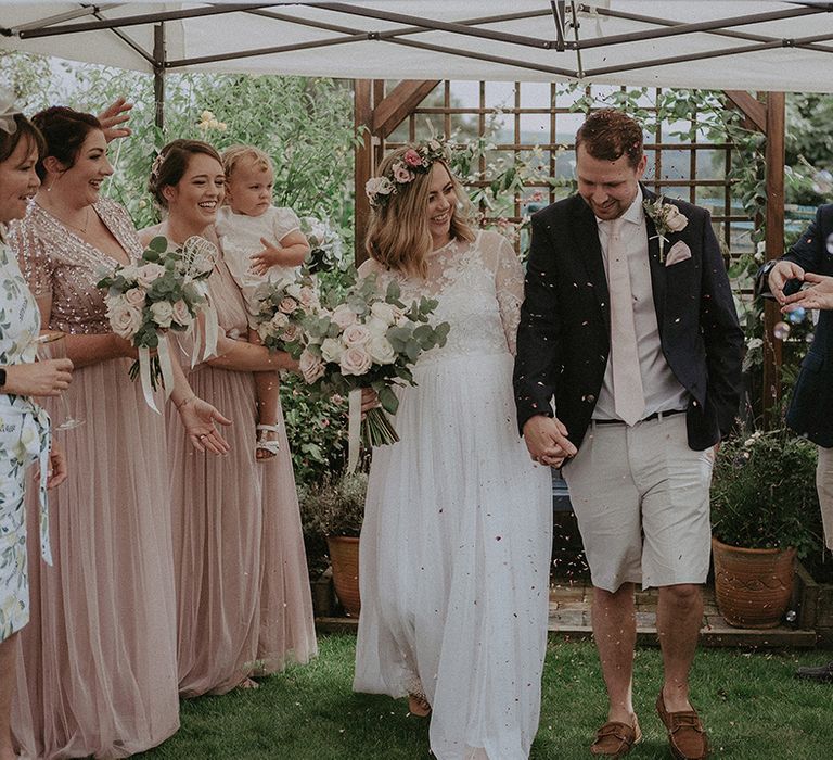 Back garden wedding confetti moment with pregnant bride in an empire line wedding dress and flower crown and groom in beige shorts and navy blazer 