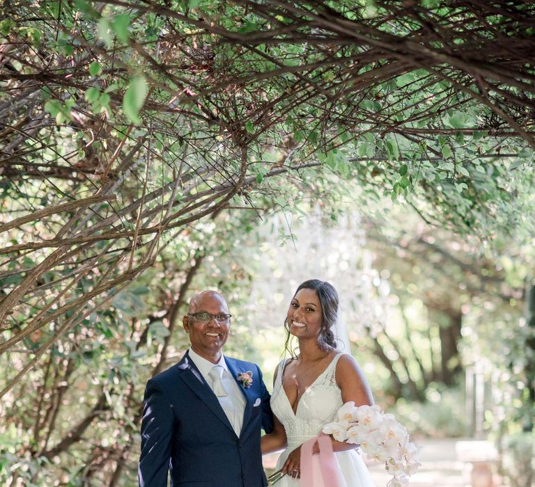 Bride stands with her father under floral archway whilst she holds white orchid bridal bouquet