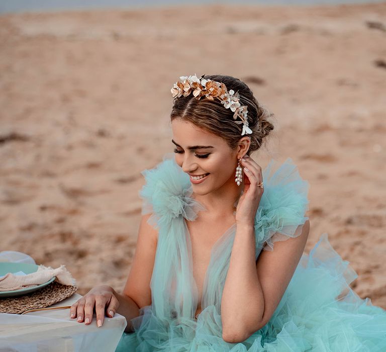 Bride in a mint green wedding dress, gold headband and dangly earrings sitting at her relaxed beach reception 