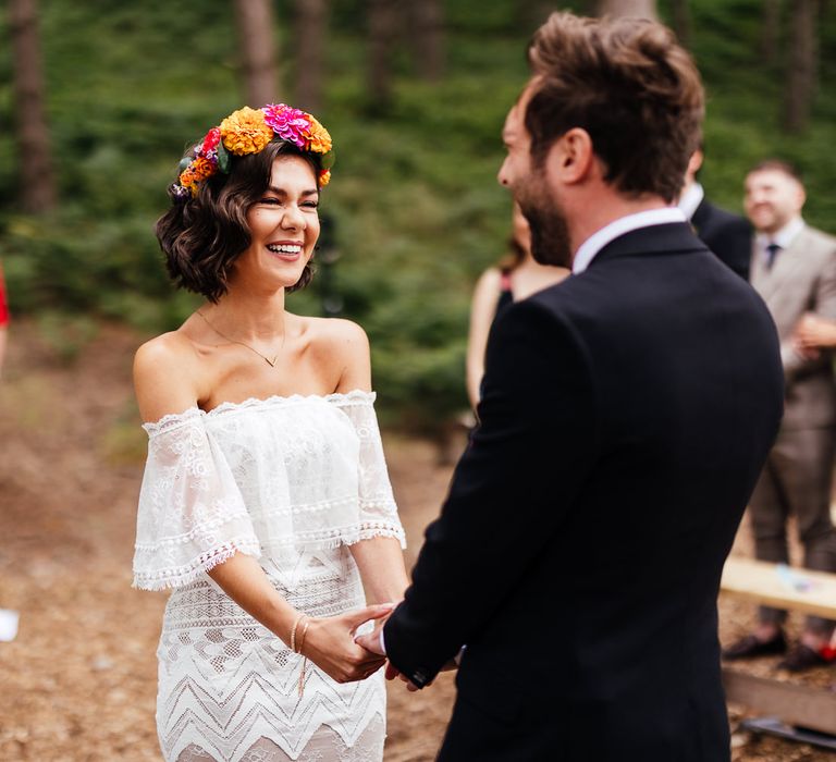 Bride in strapless Grace Loves Lace wedding dress and colourful flower crown laughs during wedding ceremony whilst holding hands with groom in black suit at outdoor wedding ceremony