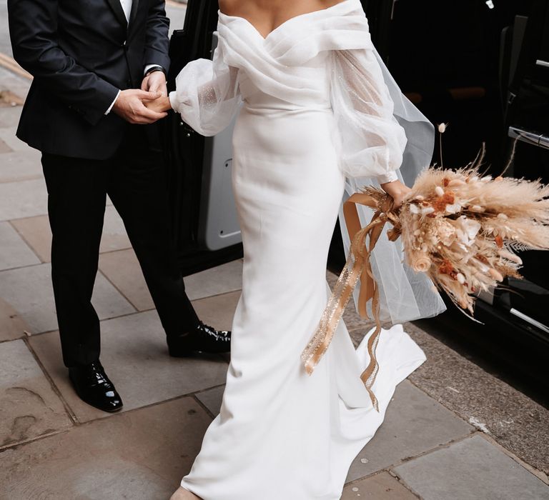 Groom in a tuxedo helping his bride out of a black cab in an off the shoulder wedding dress with long sleeves 