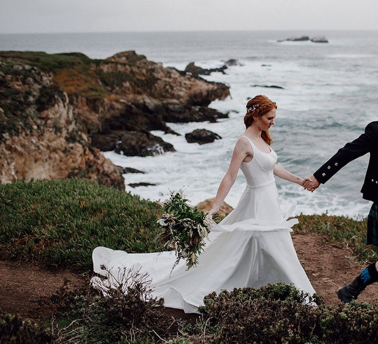 Groom in a tartan kilt holding hands with his bride in a floaty wedding dress as they walk along the cliff edges at Big Sur