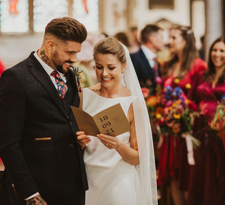 Bride in a slip wedding dress and cathedral length veil and groom in a three-piece wool suit holding a craft card order of service at their church wedding ceremony 