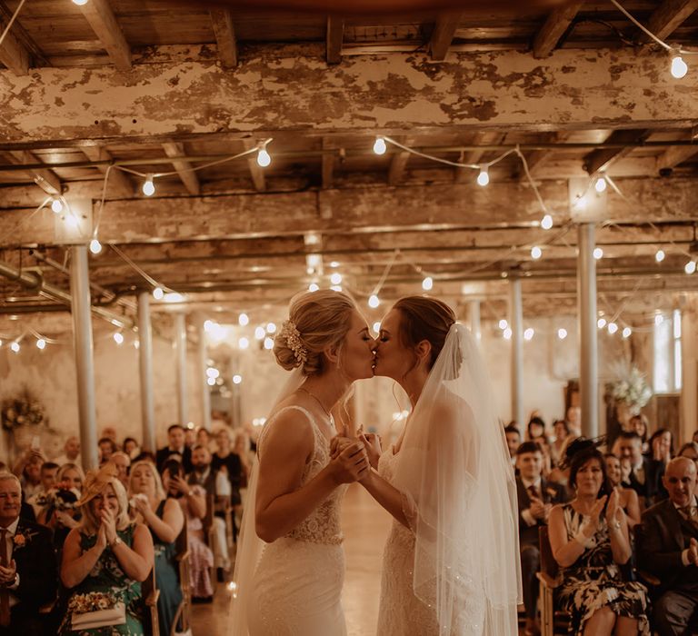 Brides kiss on their wedding day as lights hang from the ceiling 