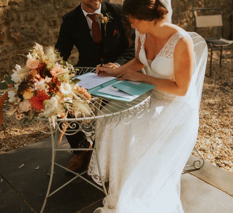 Bride in lace wedding dress and veil sits signing marriage wedding register at bistro table with groom in dark suit during outdoor wedding ceremony