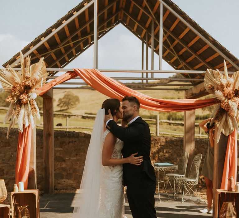 Bride in lace wedding dress with train and veil kisses groom in dark suit after outdoor wedding ceremony with rustic decor and orange draping