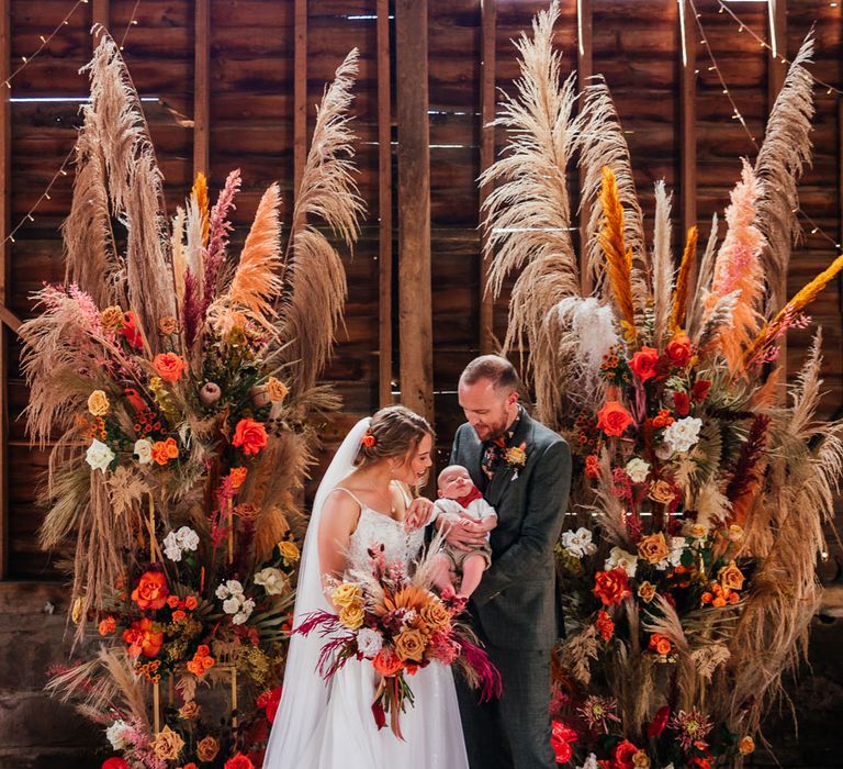 Bride in lace wedding dress with train and veil holding mixed bridal bouquet with orange florals stands with groom in green three peeve wedding suit as he holds their baby for rustic barn festival style wedding 