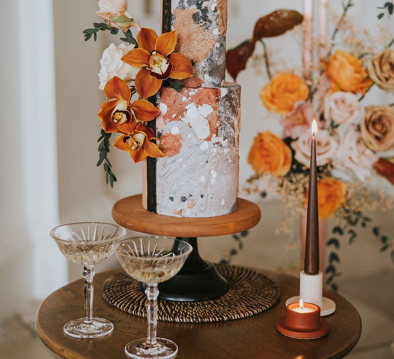 Two tier wedding cake with orange, black and white marble design on a wood and black cake stand decorated with tropical flowers 