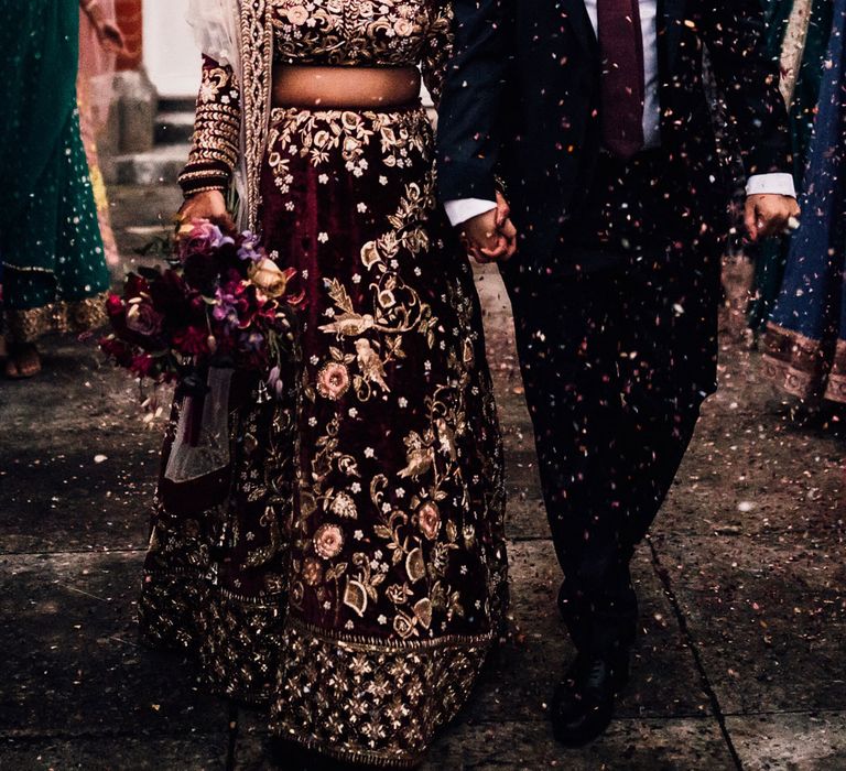 Bride in burgundy Indian saree wedding dress walks out of wedding with groom in dark suit and burgundy tie as guests throw confetti 