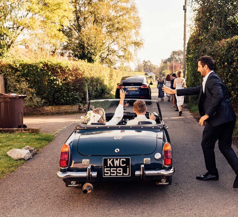 Wedding guest in black suit runs alongside bride and groom sat in vintage teal convertible 