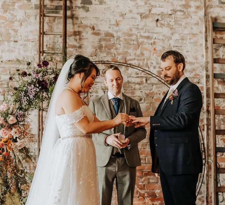 Bride in floral lace wedding dress and veil puts ring on hand of groom in blue three piece suit and brown shoes in front of floral moongate during wedding ceremony