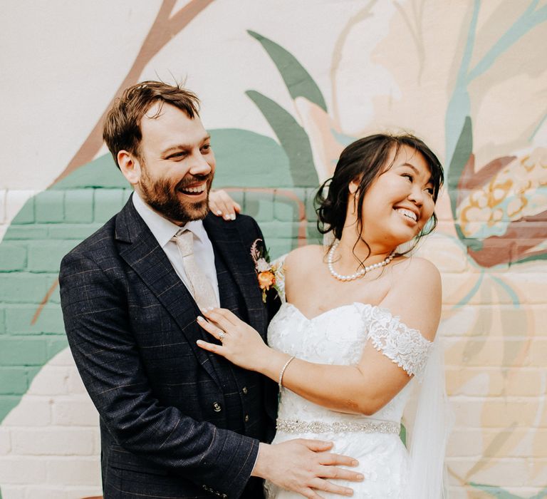 Groom in three piece check suit with light pink tie stands with bride in sweetheart neckline lace wedding dress and pearl necklace