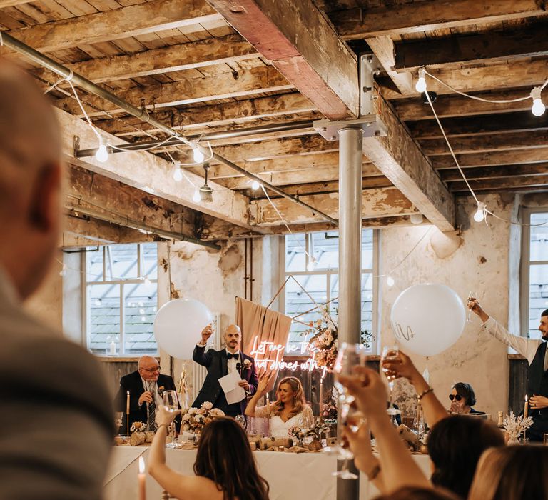 Guests raise a glass during wedding reception in industrial mill wedding venue with rustic styling and neon wedding sign