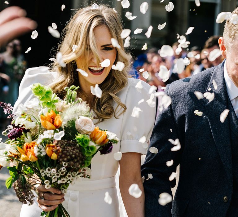 Bride holding orange bouquet and groom in navy suit jacket and tie walk through confetti