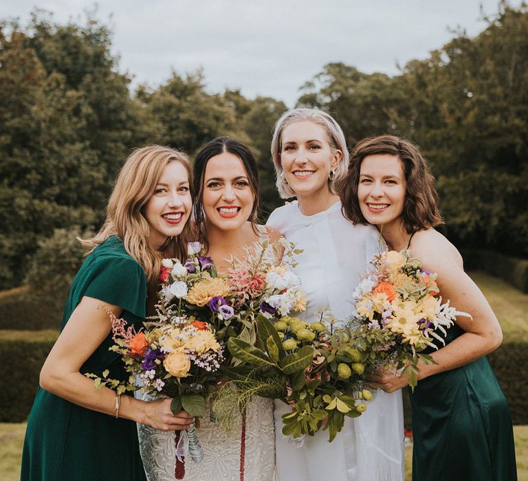 Brides pose with their two bridesmaids in green mismatched style dresses 