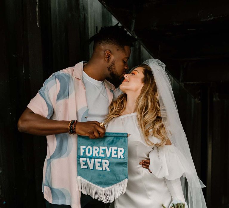 Groom in a pastel patterned shirt holding a Forever Ever teal suede fabric wedding banner 