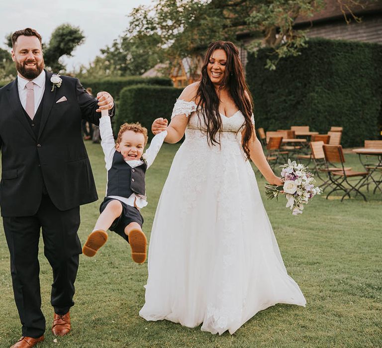 Bride and groom swing young page boy between them after their outdoor wedding ceremony at Micklefield Hall 